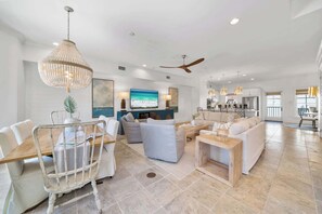 A spacious living room with a large TV, comfortable seating, a ceiling fan, and a view of the kitchen.