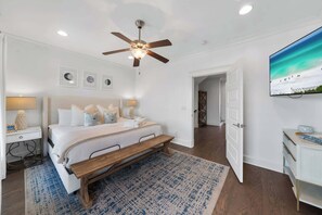 Un dormitorio con cama, mesitas de noche, ventilador de techo, televisor y una puerta que da a otra habitación.