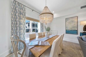 A dining area with a wooden table, white chairs, a chandelier, and a view of the outside through large windows. - New Downtime Prominence 30A Lux, Pet Friendly, LSV, Bikes (Panama City Beach)