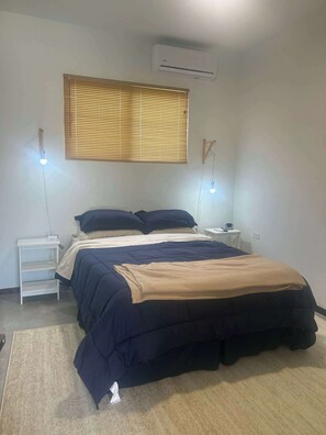 Ein Schlafzimmer mit Bett, Nachttischen, Fenster mit Jalousien und einer Klimaanlage.