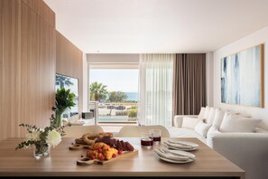 A modern hotel room with a wooden dining table set with pastries, a fruit platter, and glasses of juice. A white sofa and a painting are visible in the room. - 1-Bedroom Condo with Sea View & Pool (Voula)