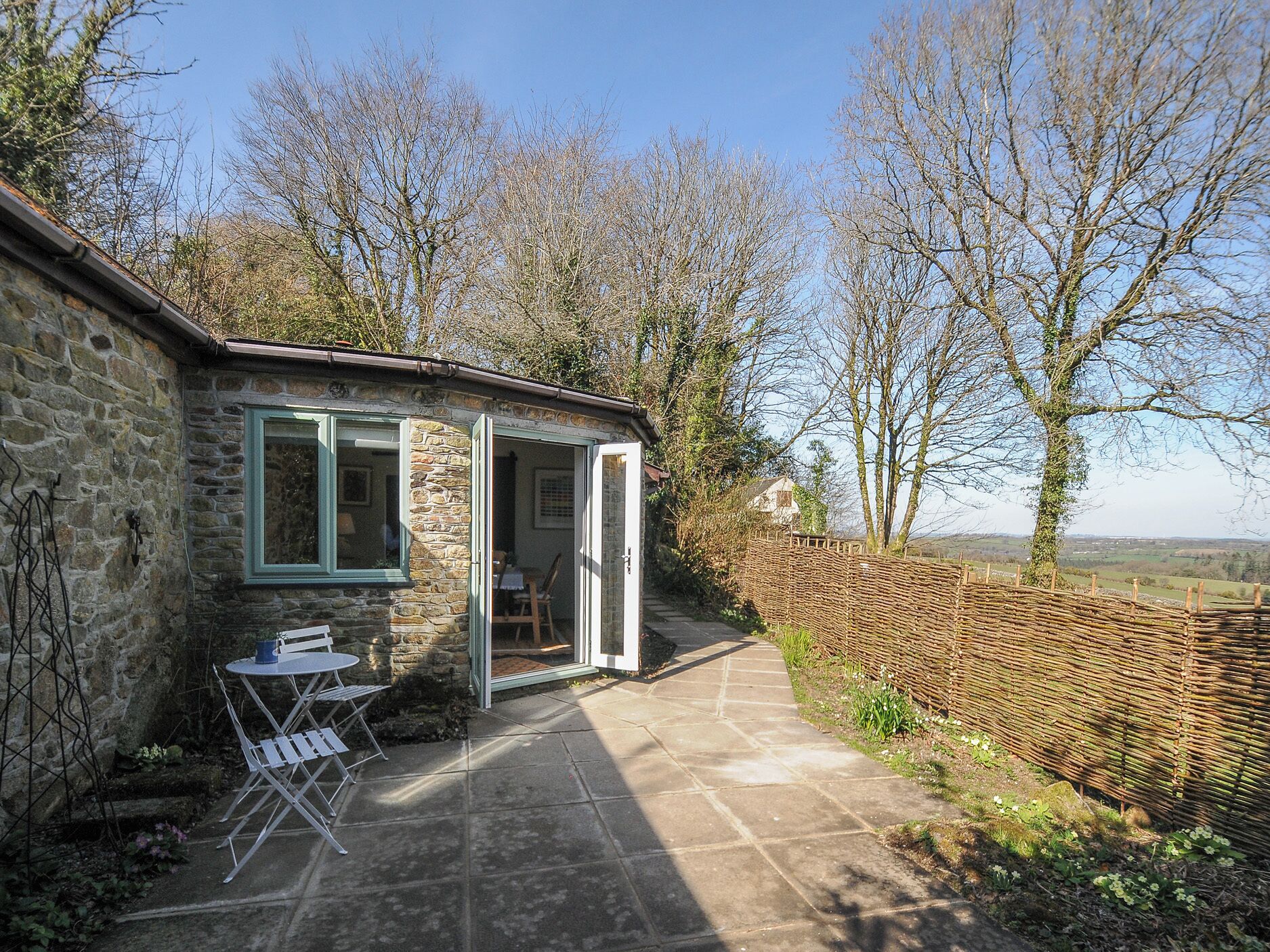 A stone cottage with a small patio, a table and chairs, and a view of the countryside.