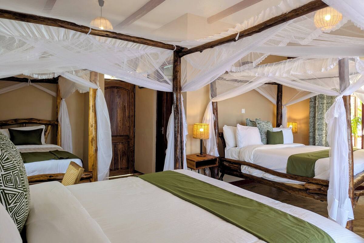 Habitación con dos camas, cada una con estructura de dosel y ropa de cama blanca. Se observa una puerta de madera y una mesita de noche con una lámpara.
