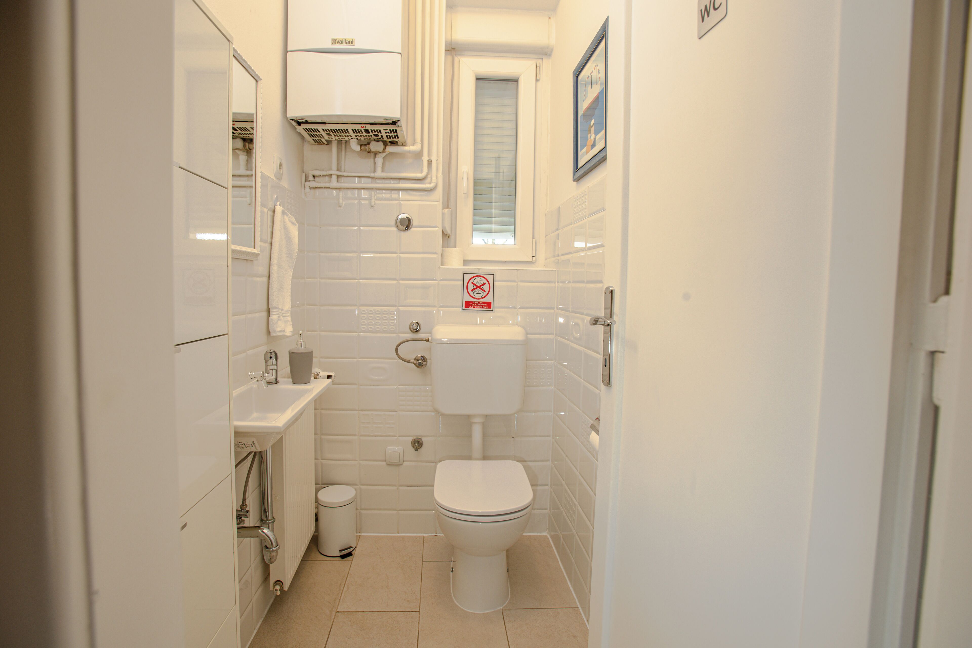 A small, clean bathroom with white tiles, a toilet, a sink, and a window with blinds.