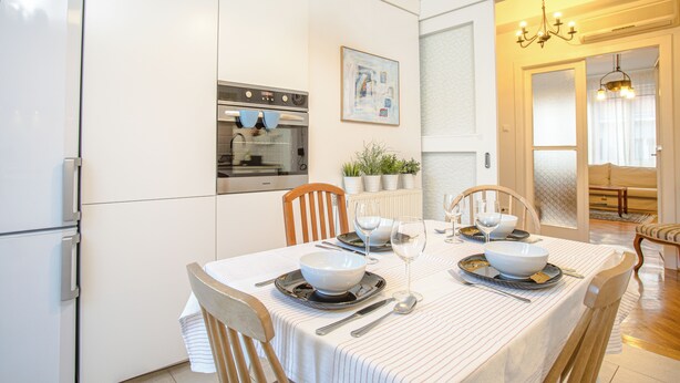 A modern kitchen with white cabinetry, a built-in oven, and a dining area set for four with a striped tablecloth, white bowls, and wine glasses.