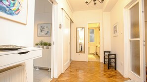 A hallway with a wooden floor, white walls, and a ceiling light. There are doors leading to other rooms, and a small table with a chair in the corner.