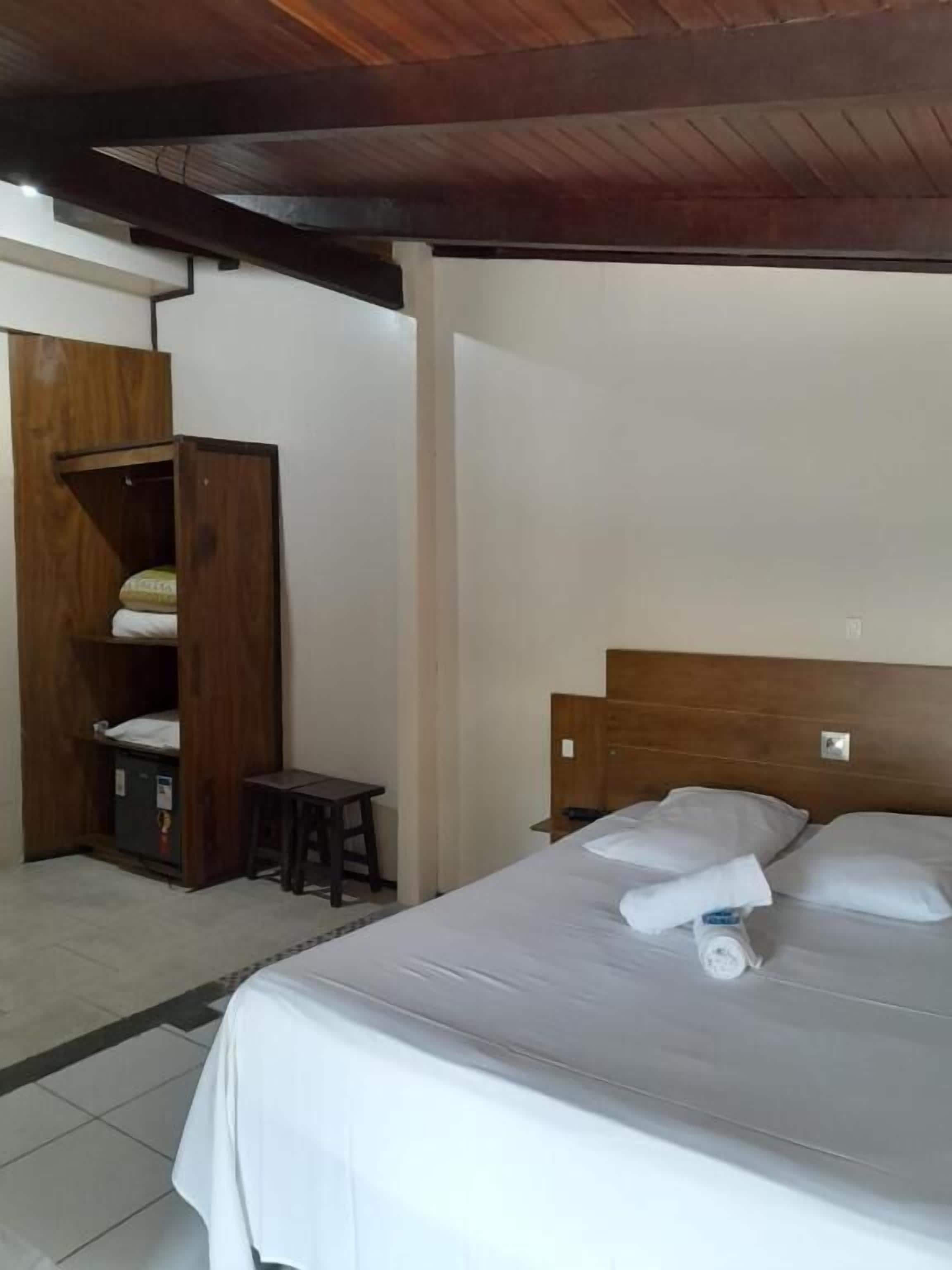 Een hotelkamer met een houten hoofdeinde, een bed met witte lakens, een klein houten tafeltje en een kledingkast.