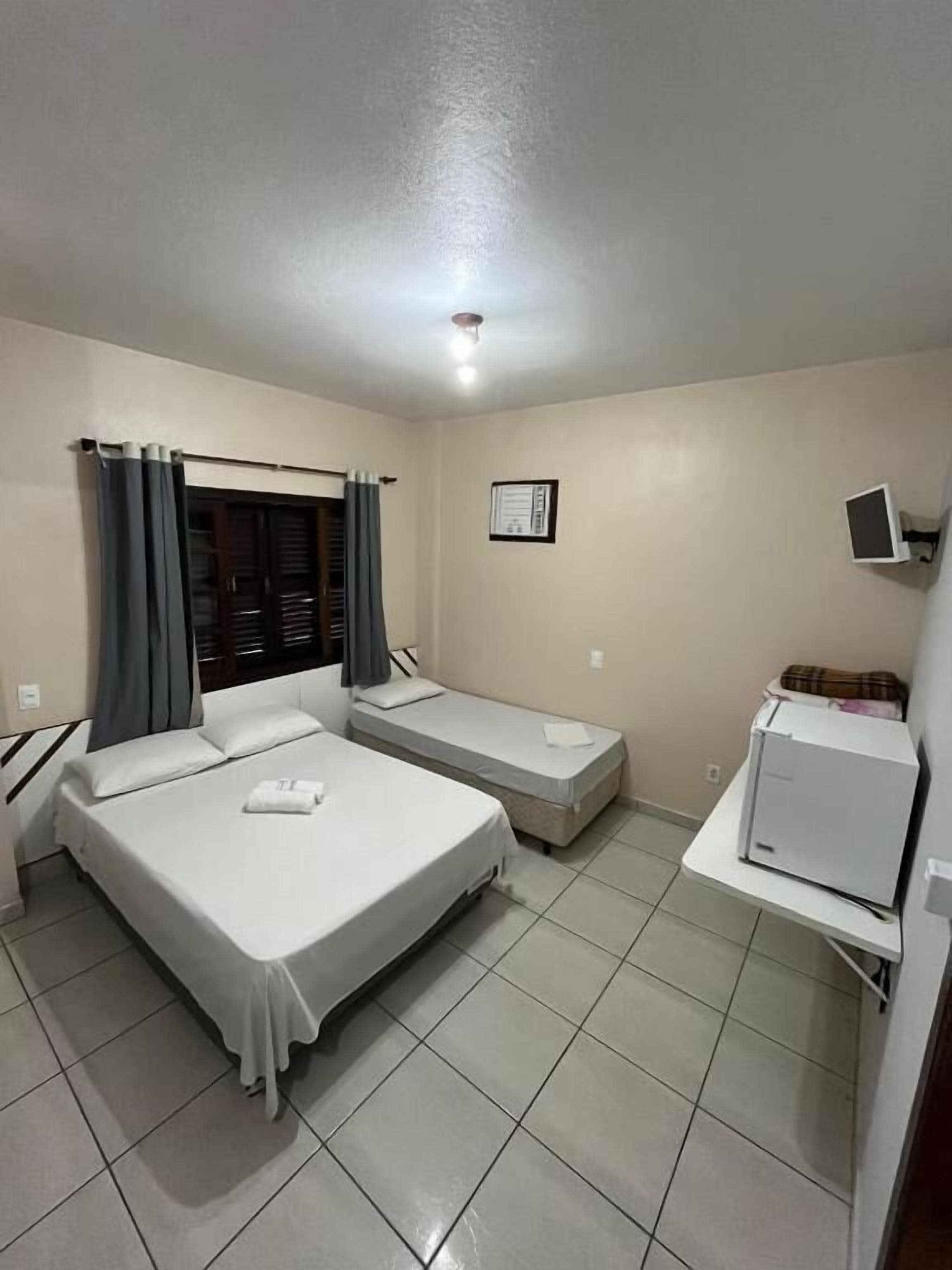 Quarto de hotel com duas camas, uma pequena mesa, um frigorífico e uma janela com cortinas.