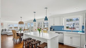A spacious kitchen with white cabinets, a large island with stools, and blue pendant lights. - Ocean Views! Walk to Beaches! 4BR Vacation Retreat (Kennebunk)