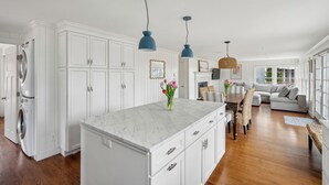 A spacious kitchen with white cabinetry, a marble island, and a dining area with a gray sofa and wicker chairs. - Ocean Views! Walk to Beaches! 4BR Vacation Retreat (Kennebunk)