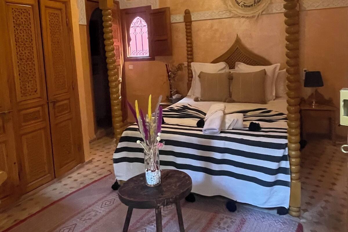 A bedroom with a four-poster bed, a chandelier, a wooden wardrobe, and a small table with a vase.