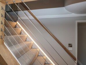 A modern staircase with wooden handrails and a metal railing, leading to a room with a door and a light switch on the wall. - Dove Cottage (Ambleside)