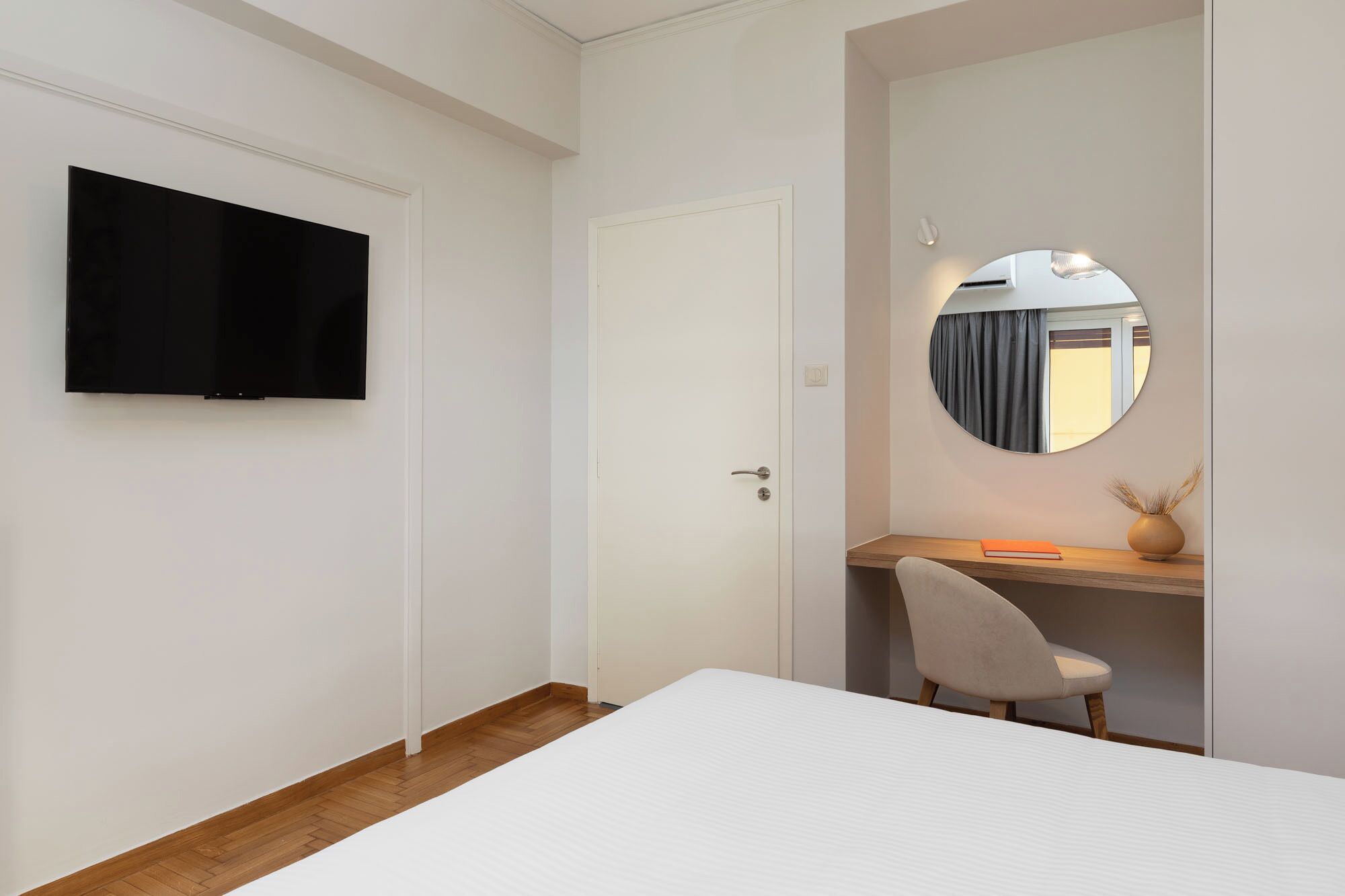 Une chambre à coucher avec un lit, un bureau et une chaise, un téléviseur fixé au mur et un miroir rond également fixé au mur.