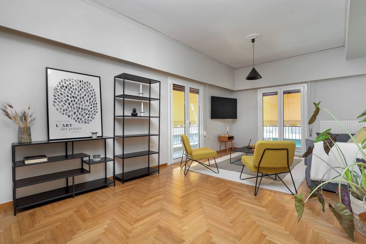 A modern living room with a herringbone wooden floor, a black bookshelf, a yellow armchair, a black sofa, and a flat-screen TV mounted on the wall.