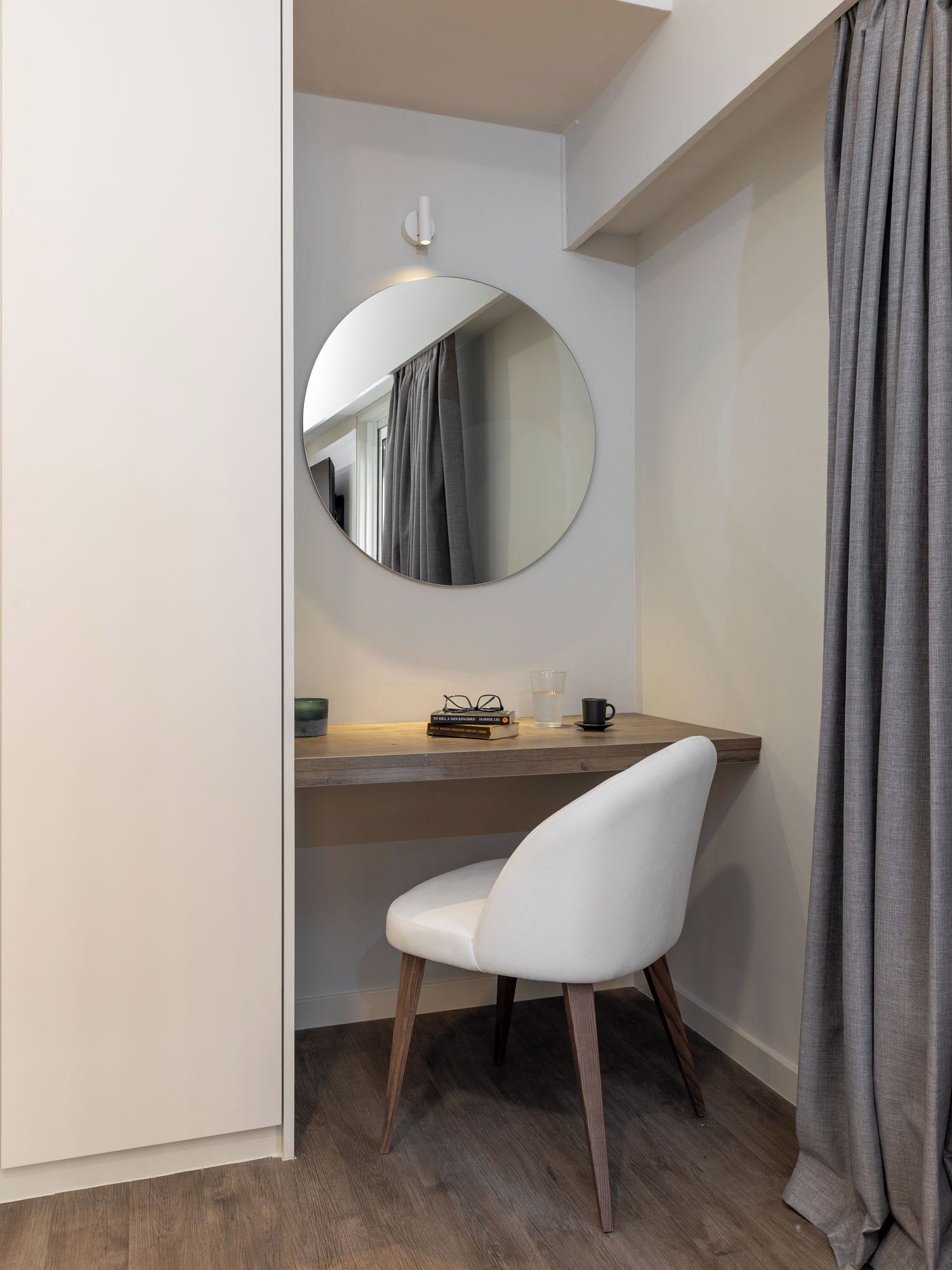 A small, modern room with a round mirror, a white chair, a wooden desk, and a grey curtain.