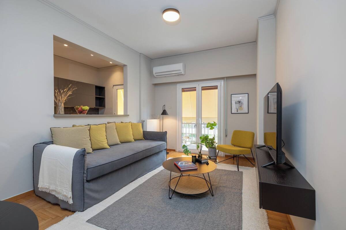 A modern living room with a grey sofa, yellow cushions, a round coffee table, and a flat-screen TV.