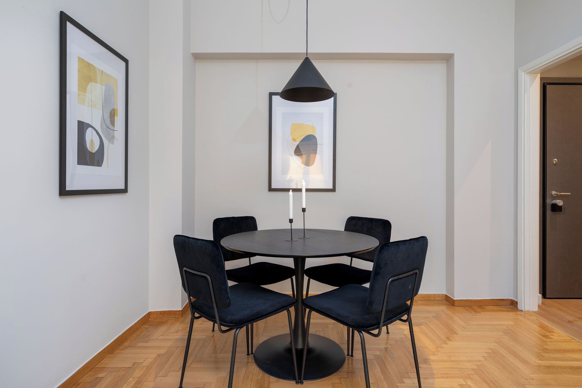 A dining area with a round table, four black chairs, a black pendant light, and two framed artworks on the wall.