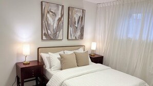 Una cama bien hecha con colcha blanca, dos lámparas de noche y una mesita de noche con lámpara. Se observan dos cuadros abstractos en la pared sobre la cama.