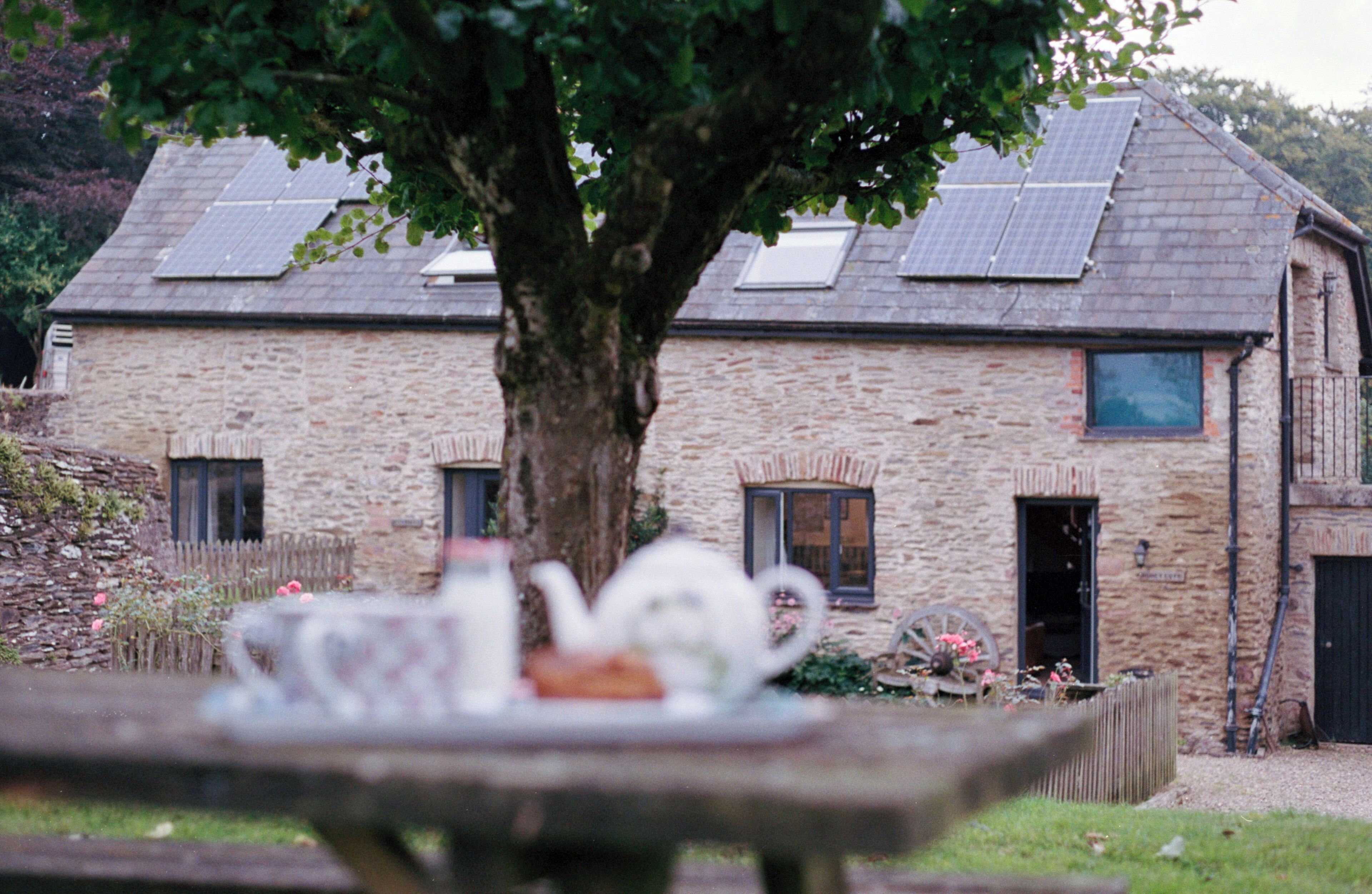A stone building with solar panels, a tree in front, and a table with a teapot and cups.