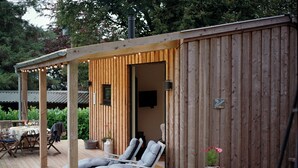 Une terrasse en bois avec deux chaises longues et une table, un espace salon couvert, et un bâtiment en bois sur lequel est apposée une enseigne indiquant « Mokden ».