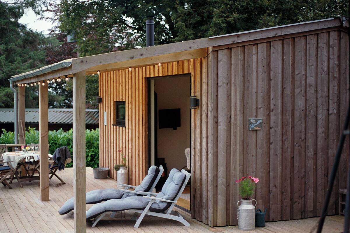 A wooden deck with two lounge chairs and a table, a covered seating area, and a wooden building with a sign that reads "Mokden."
