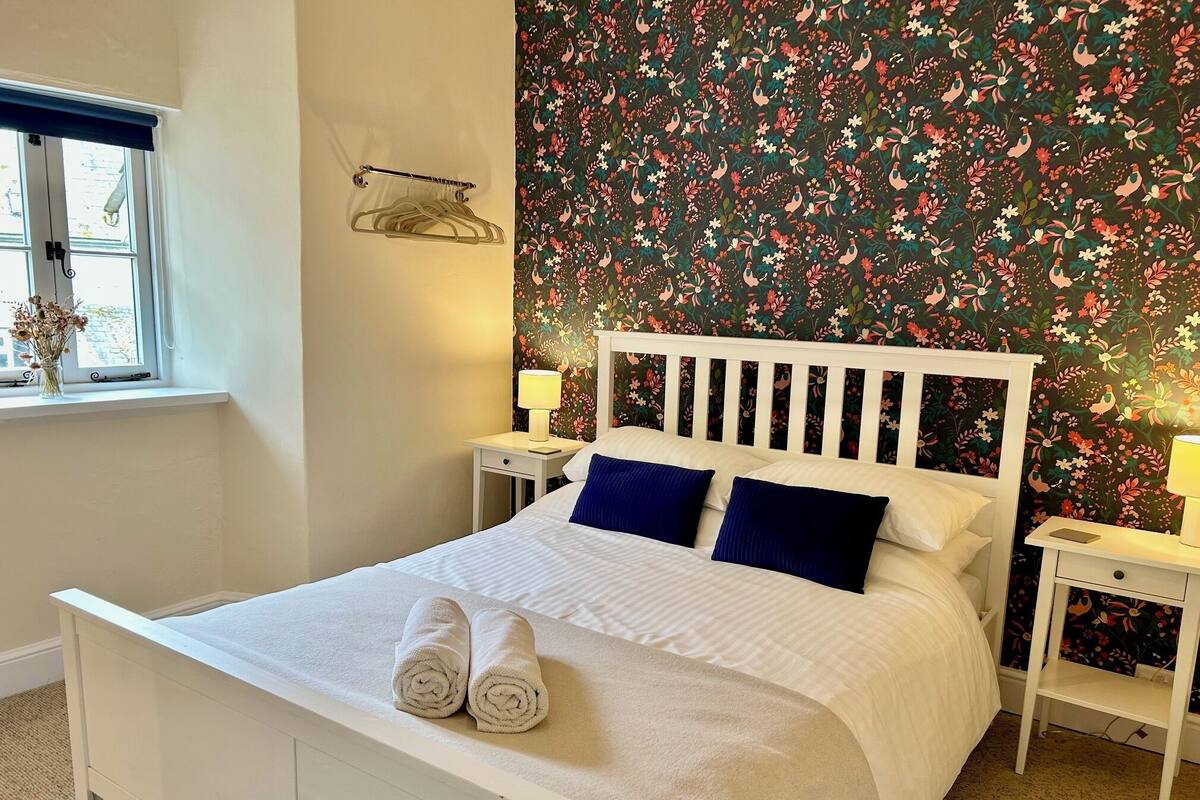 A neatly made bed with white linens and two blue pillows, a window with a blue blind, a floral wallpaper, and a small bedside table with a lamp.