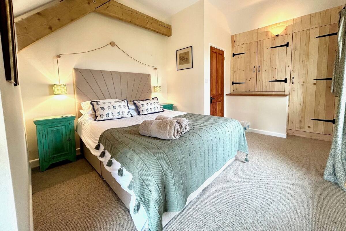 A bedroom with a bed, a nightstand, a wooden wardrobe, and a ceiling with exposed beams.