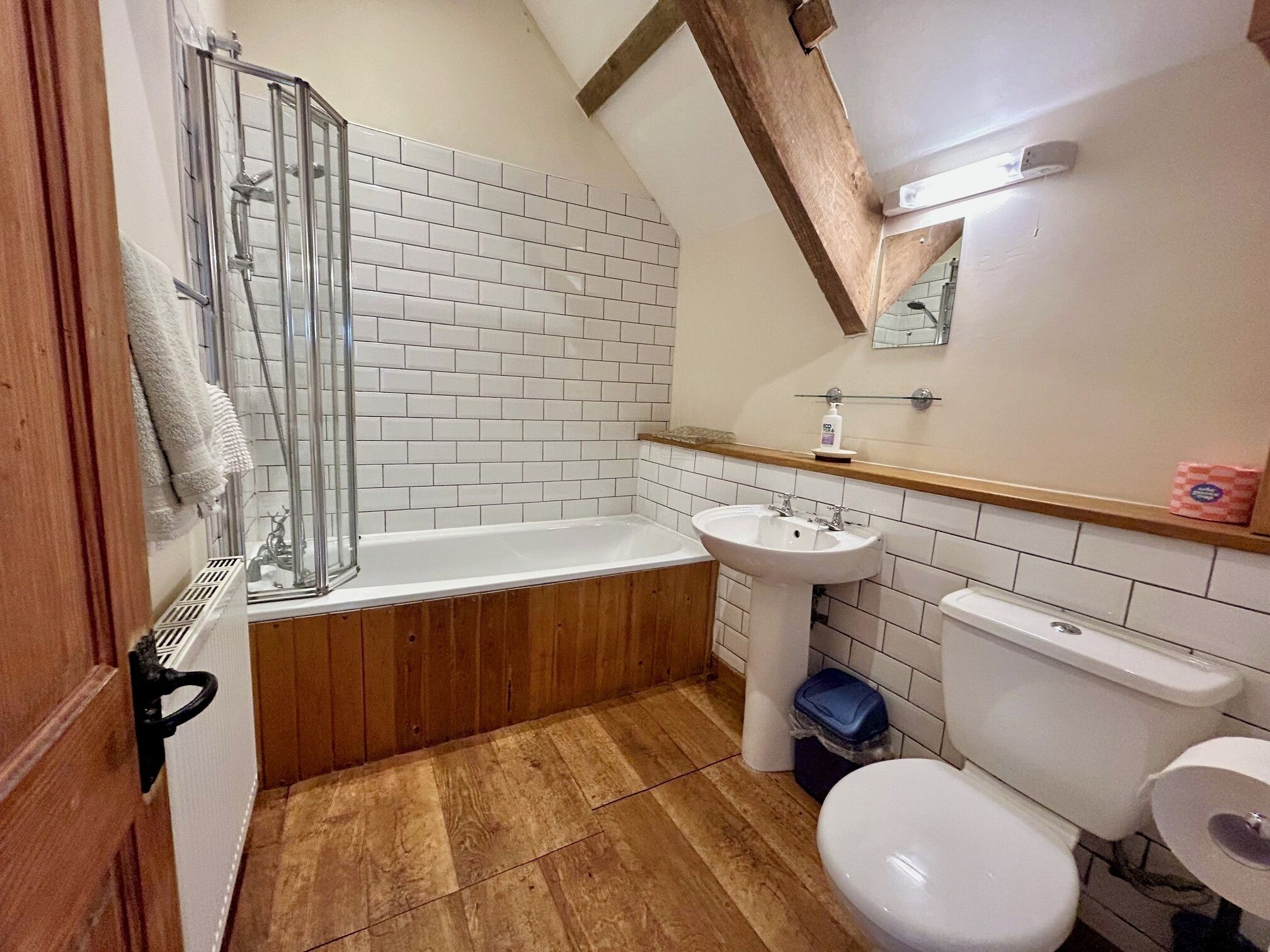 A bathroom with a shower, sink, toilet, and wooden flooring.