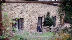 Un bâtiment en pierre avec deux fenêtres et une porte, entouré de verdure et d’une clôture en bois.