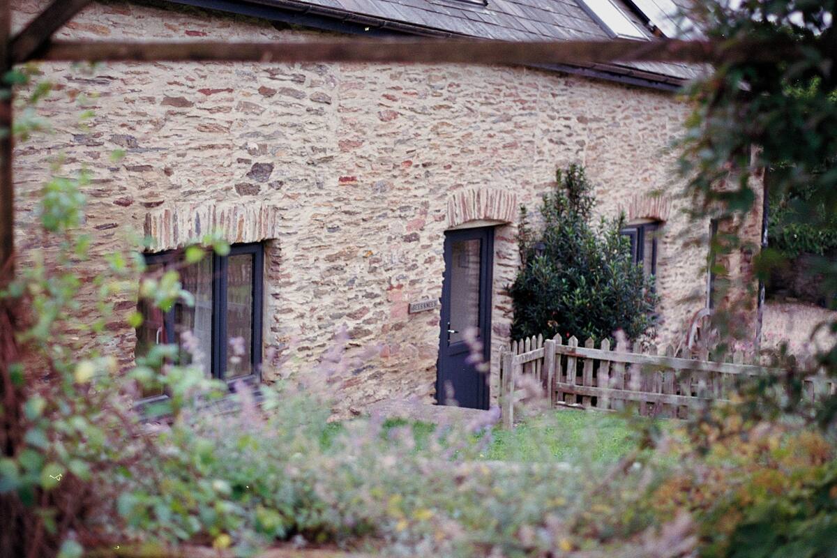 A stone building with two windows and a door, surrounded by greenery and a wooden fence.