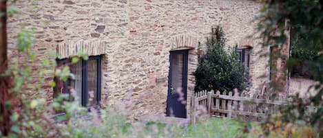 Un edificio de piedra con dos ventanas y una puerta, rodeado de follaje y una valla de madera.