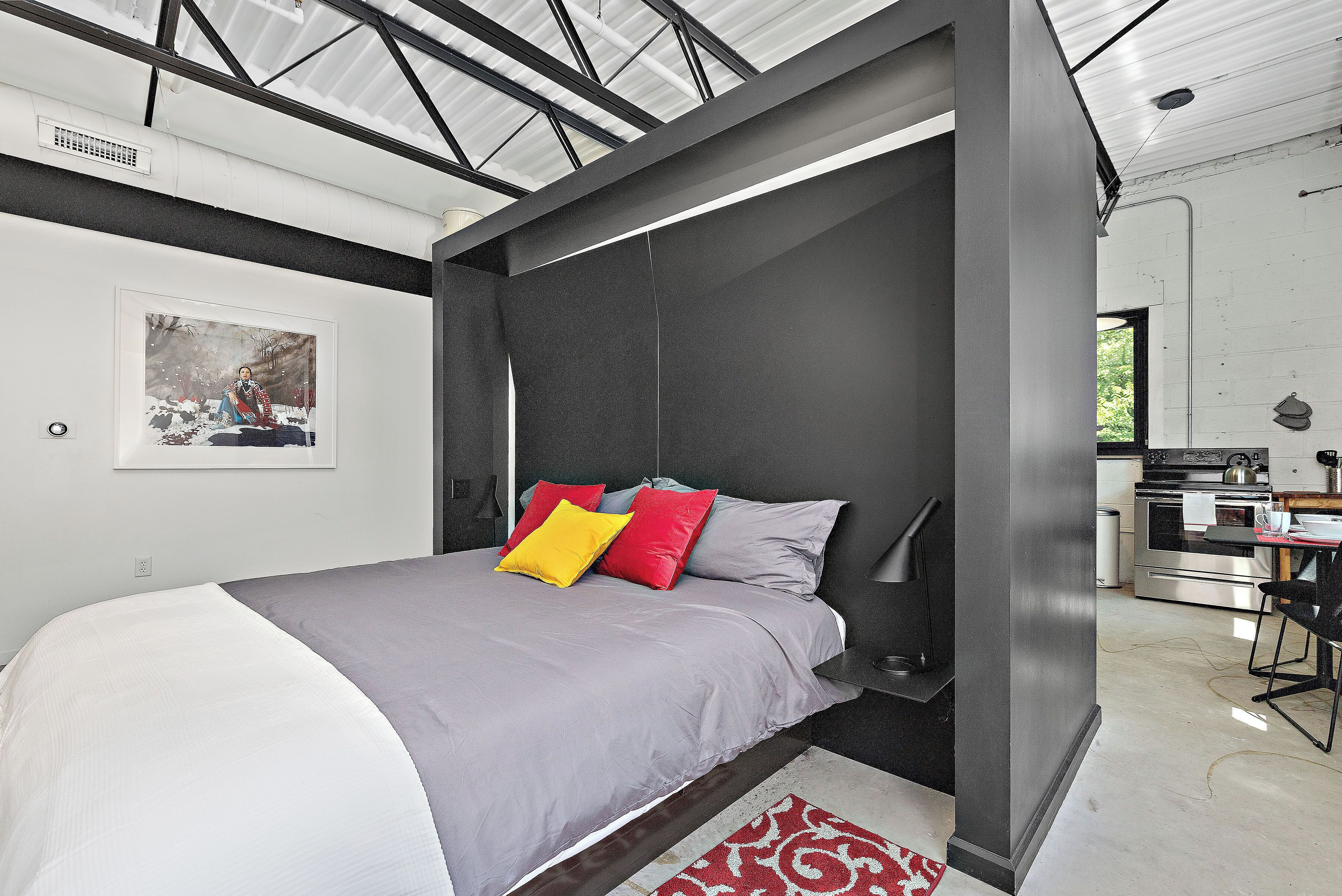 Une chambre à coucher moderne avec un lit, une table de chevet et un coin cuisine visible à travers une porte ouverte.