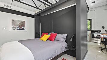 Une chambre à coucher moderne avec un lit, une table de chevet et un coin cuisine visible à travers une porte ouverte.