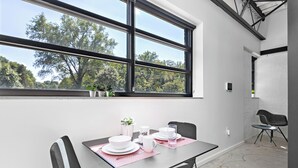 A dining area with a table set for two, a window with a view of trees, and a chair in the corner.