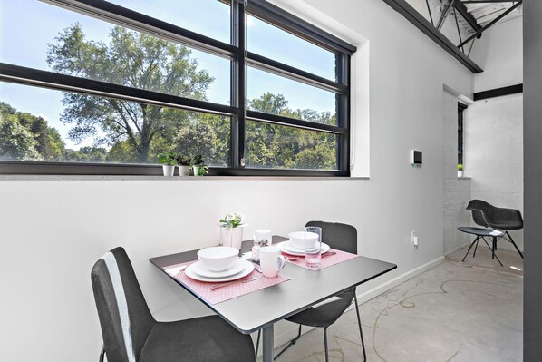 Un comedor con una mesa dispuesta para dos, una ventana con vista a árboles y una silla en la esquina.