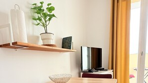 Ein kleiner, gut beleuchteter Raum mit einem Holztisch, einem Stuhl, einem Fernseher, einer Topfpflanze und einem Fenster mit Vorhängen.