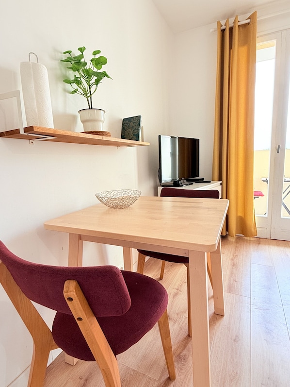 A small, well-lit room with a wooden table, a single chair, a television, a potted plant, and a window with curtains.