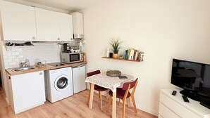 A compact kitchen with a washing machine, a microwave, and a coffee maker. There's a dining table with chairs and a small TV on a dresser.