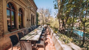 Un espace de restauration en plein air avec une longue table et des chaises, un bâtiment en pierre doté de fenêtres en arc de cercle et une vue sur une piscine.