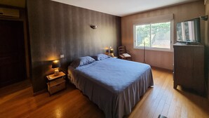 Un dormitorio con una cama grande, una mesita de noche, una silla, un televisor y una ventana con vistas al verdor.