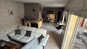 Amplia sala de estar con un sofá en forma de L, una mesa de centro de madera y una chimenea. En el fondo, hay un comedor con una araña. El piso es de piedra y las ventanas son grandes.