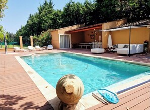 A swimming pool with a wooden deck, lounge chairs, a sofa, and a covered patio area.