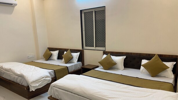 A hotel room with two single beds, each with a wooden headboard, white bedding, and decorative pillows. There is a window with a metal screen, a wall-mounted air conditioning unit, and a small table between the beds.