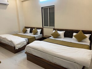 Une chambre d’hôtel avec deux lits simples, chacun doté d’une tête de lit en bois, de draps blancs et de coussins décoratifs. On y trouve une fenêtre avec un moustiquaire métallique, un climatiseur fixé au mur et une petite table entre les lits.