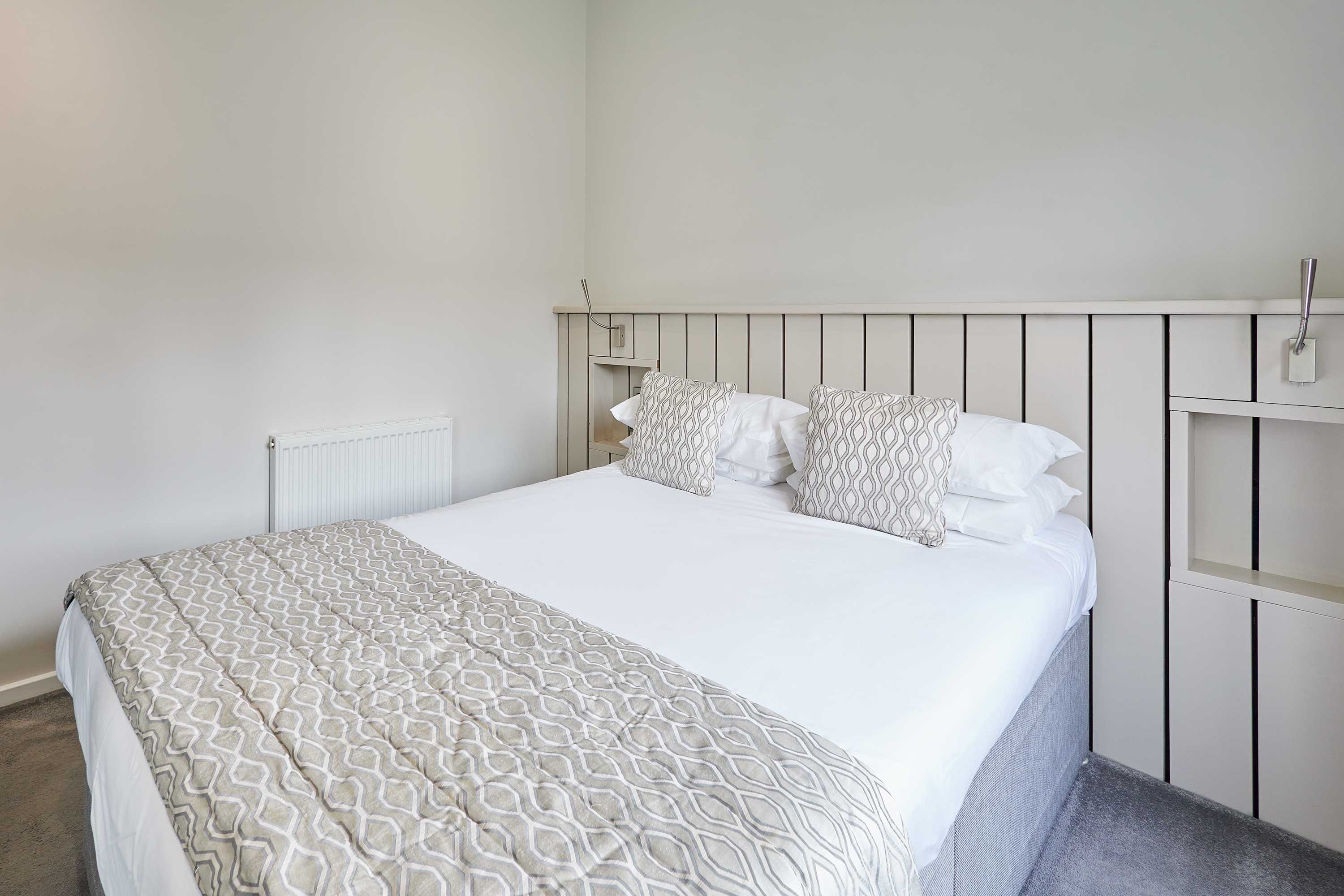 Un letto rifatto con cura, coperta e cuscini a motivi decorativi, una testiera bianca e una piccola scaffalatura a destra.