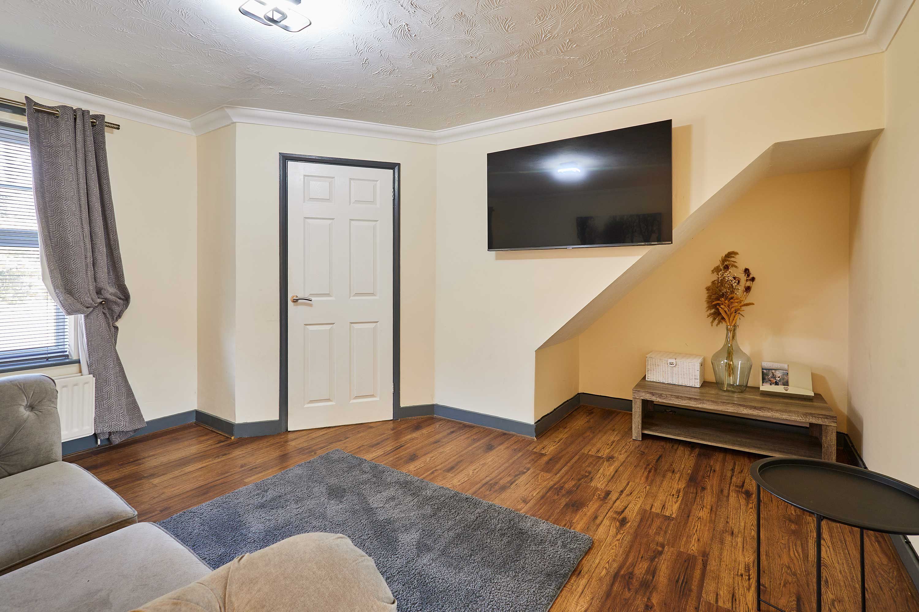 A living room with a sofa, a coffee table, a TV mounted on the wall, and a door.