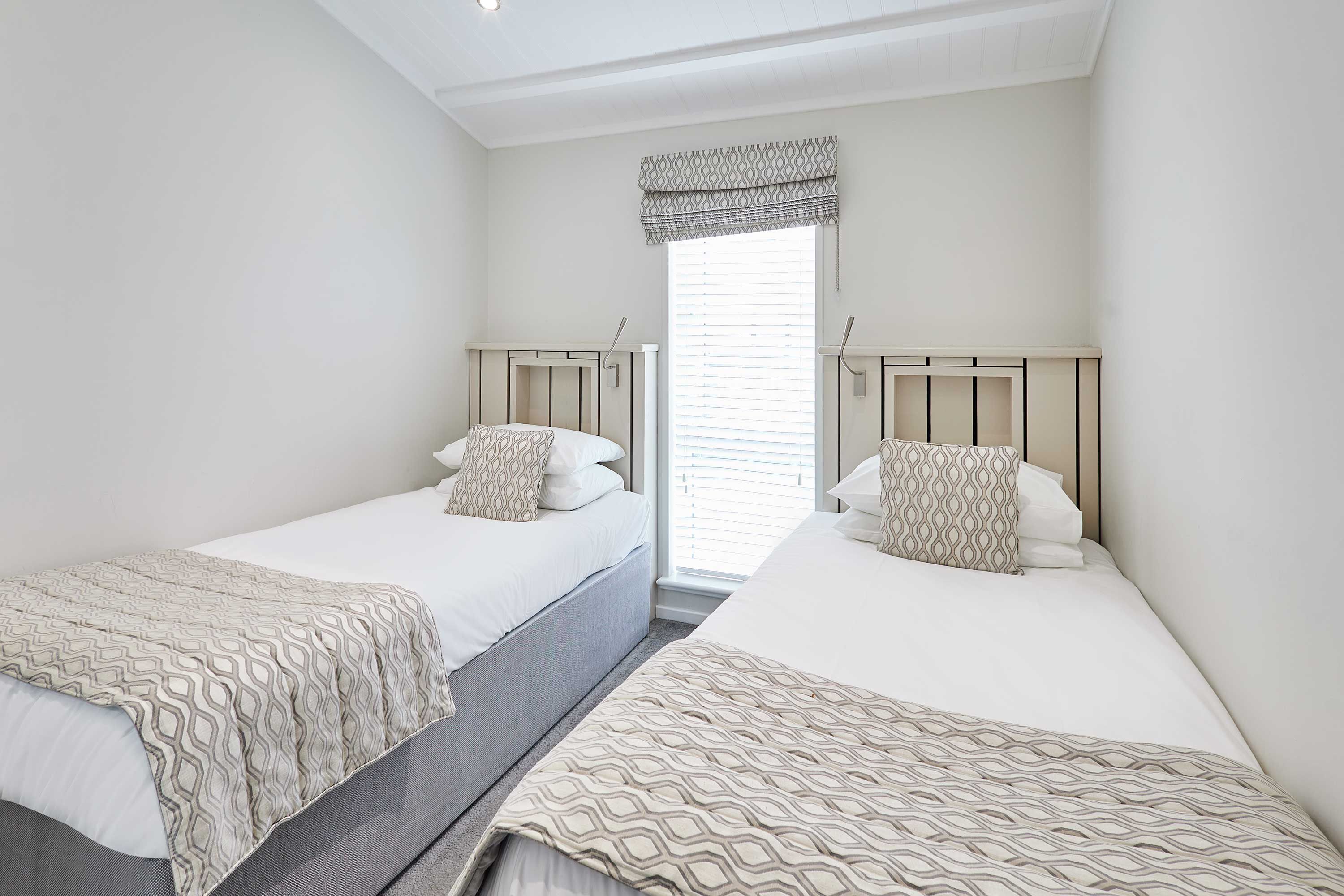 Quarto com duas camas, cada uma com roupa de cama estampada, uma janela com persianas e iluminação embutida no teto.