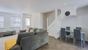 A modern living room with a grey sofa, a dining table with chairs, a staircase, and a wall clock. - Host Stay Farm Road (Hove)