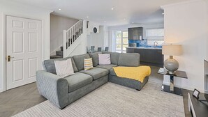 A modern living room with a grey sectional sofa, a staircase, and a kitchen in the background. - Host Stay Farm Road (Hove)