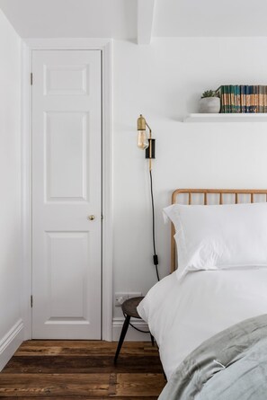 A bedroom with a white door, a bedside table with a lamp, a bed with white bedding, and a shelf with books and a plant. - Central Georgian Townhouse With Free Parking (Bath)
